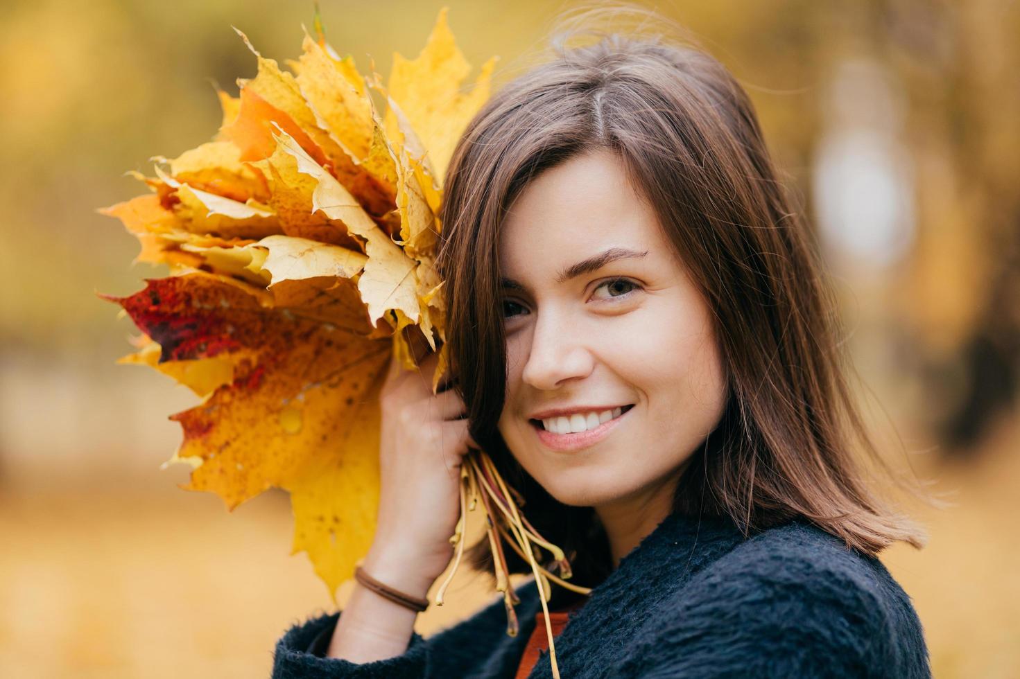 primer plano de una mujer bonita con peinado ondulado, sonrisa con dientes, disfruta del descanso durante el fin de semana, camina en el parque, lleva follaje amarillo, tiene una piel sana. concepto de personas, felicidad y recreación foto