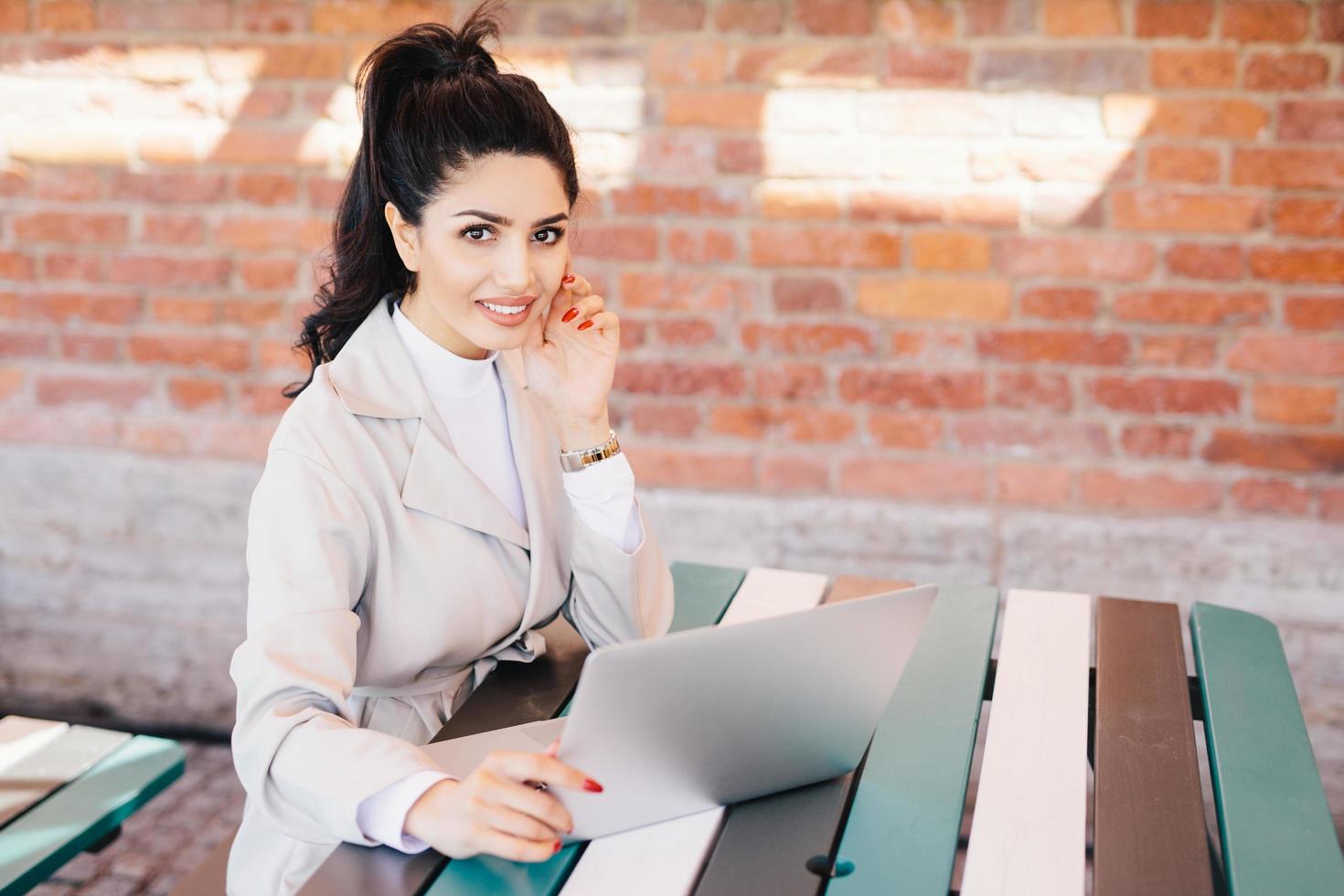 Succsessful businesswoman with dark hair, nice eyes, healthy skin, well-shaped lips having red long nails wearing formal clothes while sitting at cafe using laptop for online communication and work photo