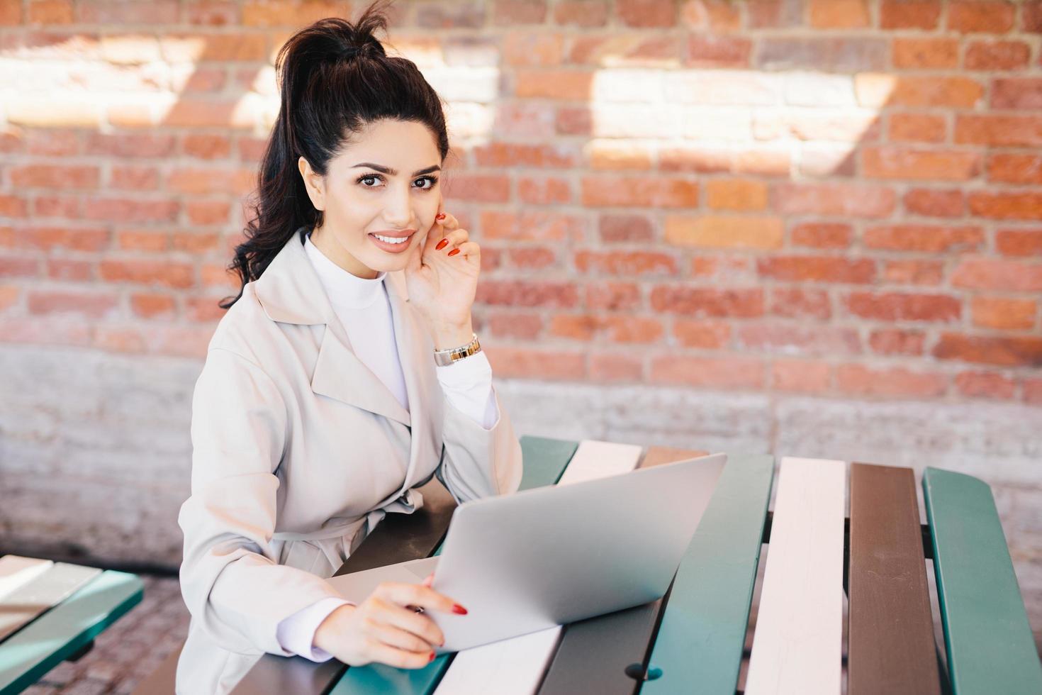 mujer de negocios exitosa con cabello oscuro, ojos bonitos, piel sana, labios bien formados con uñas largas rojas y ropa formal mientras se sienta en un café usando una computadora portátil para comunicarse y trabajar en línea foto