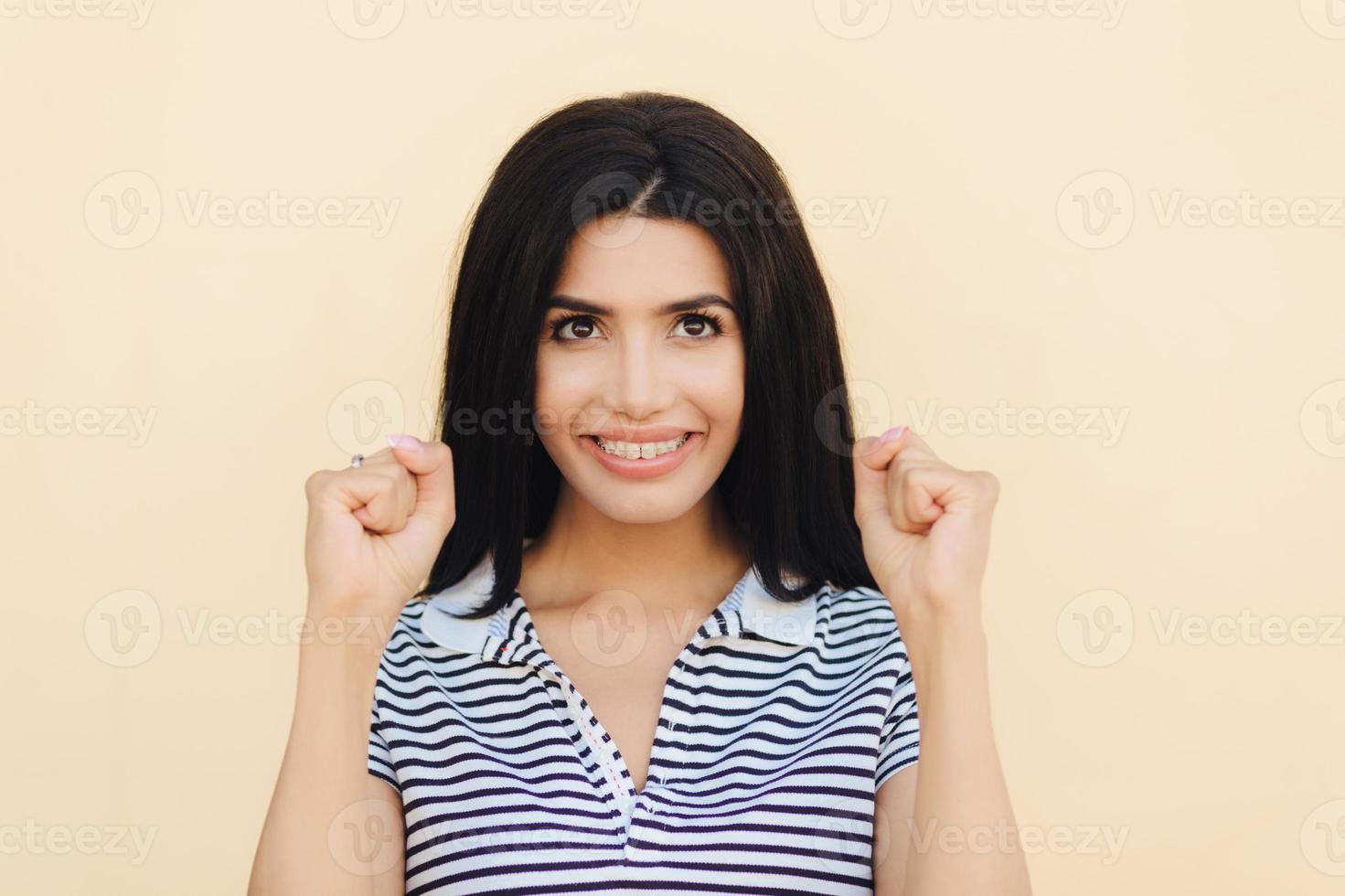 Headshot of happy brunette female model clenches fists, hopes for something good, has broad smile with white perfect teeth, celebrates success, isolated over beige studio wall. I will do it photo