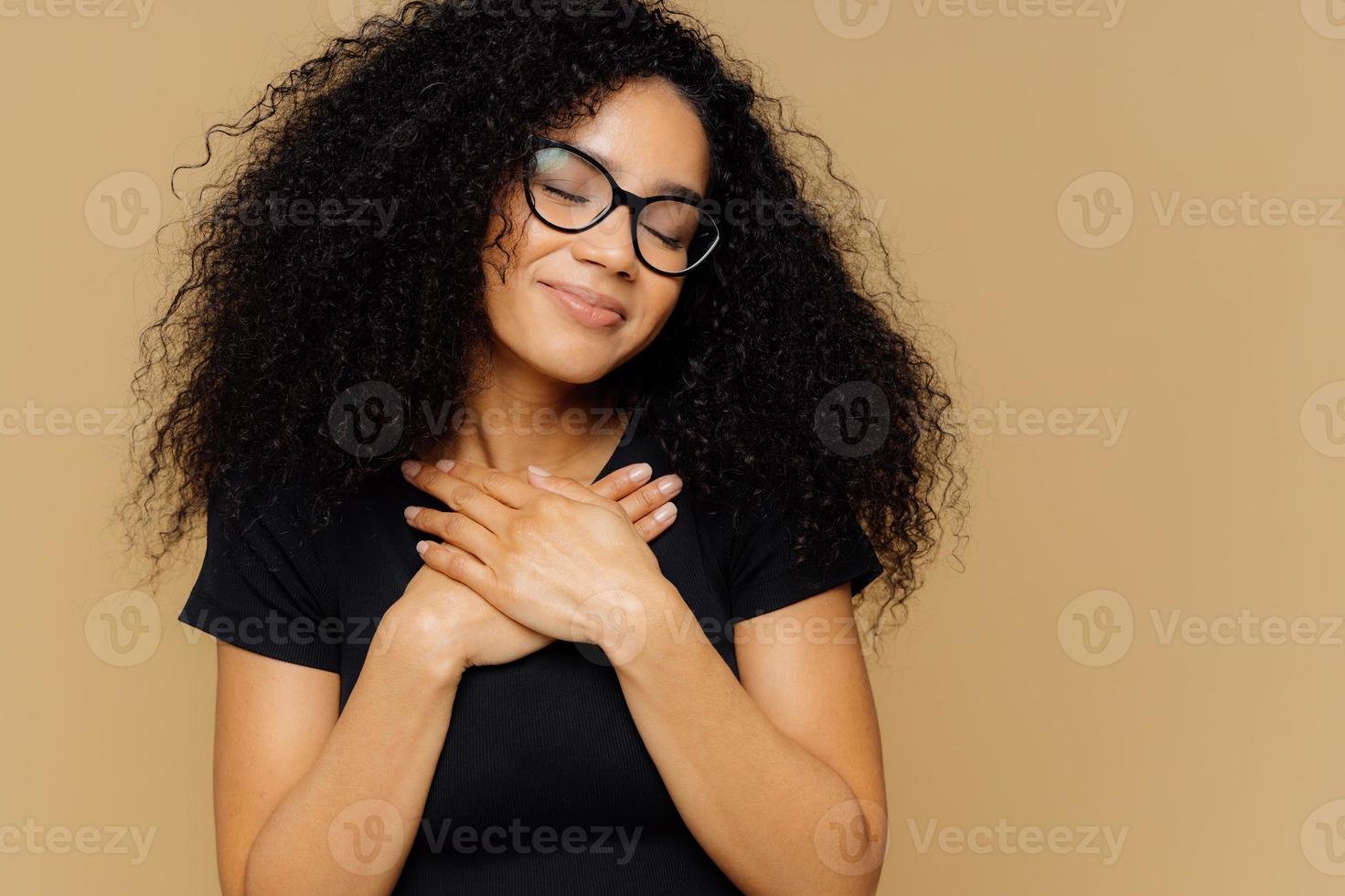 Touched lovely female with Afro hairstyle, keeps both palms on chest, has eyes shut from pleasure, hears heart piercing story, wears spectacles and casual t shirt, models against beige background photo