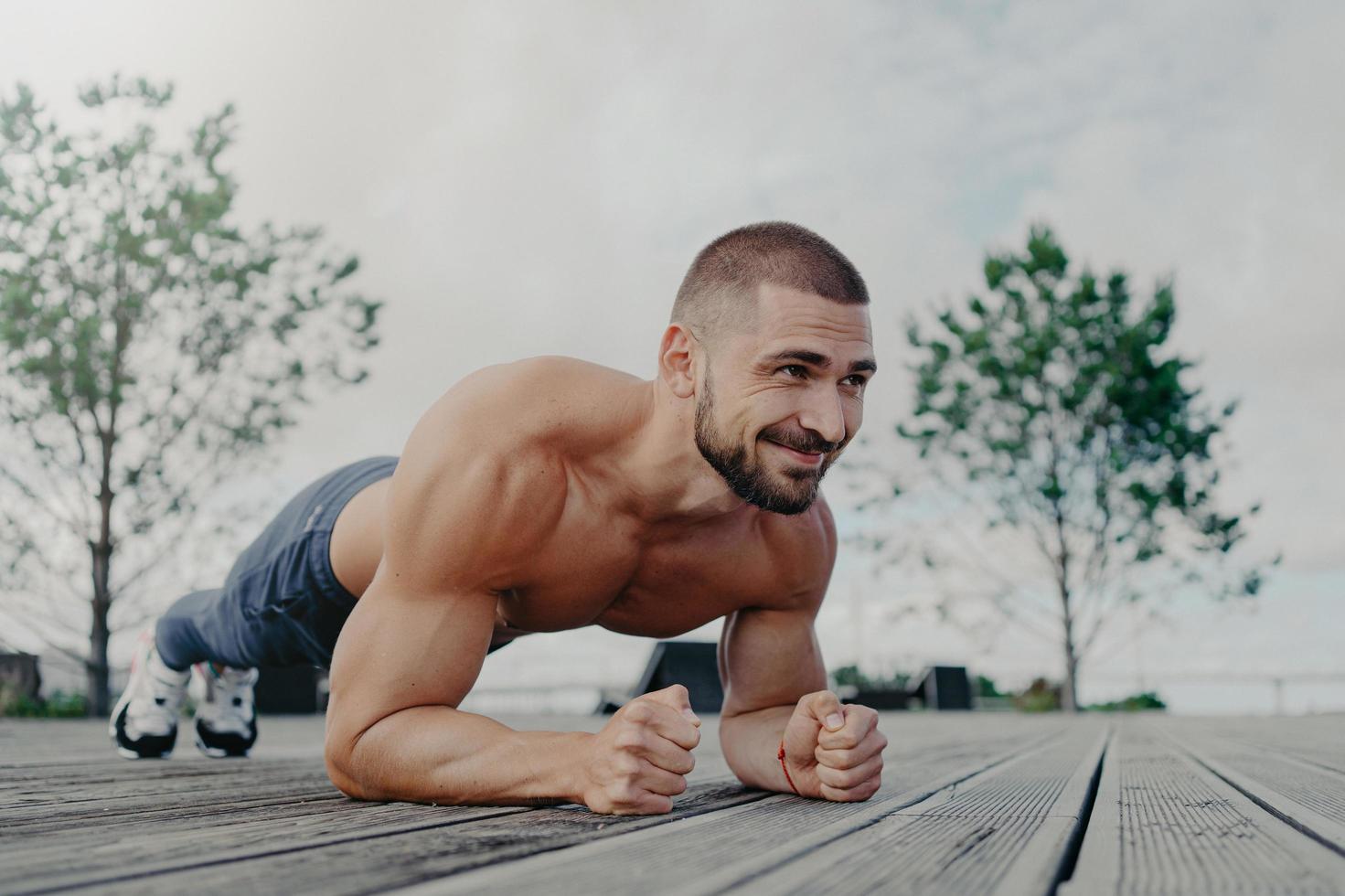 Shirtless determined bearded man with naked torso stands in plank pose and smiles gladfully, demonstrates endurance, poses outdoor, thinks about his body and health. Athletic guy trains outside photo