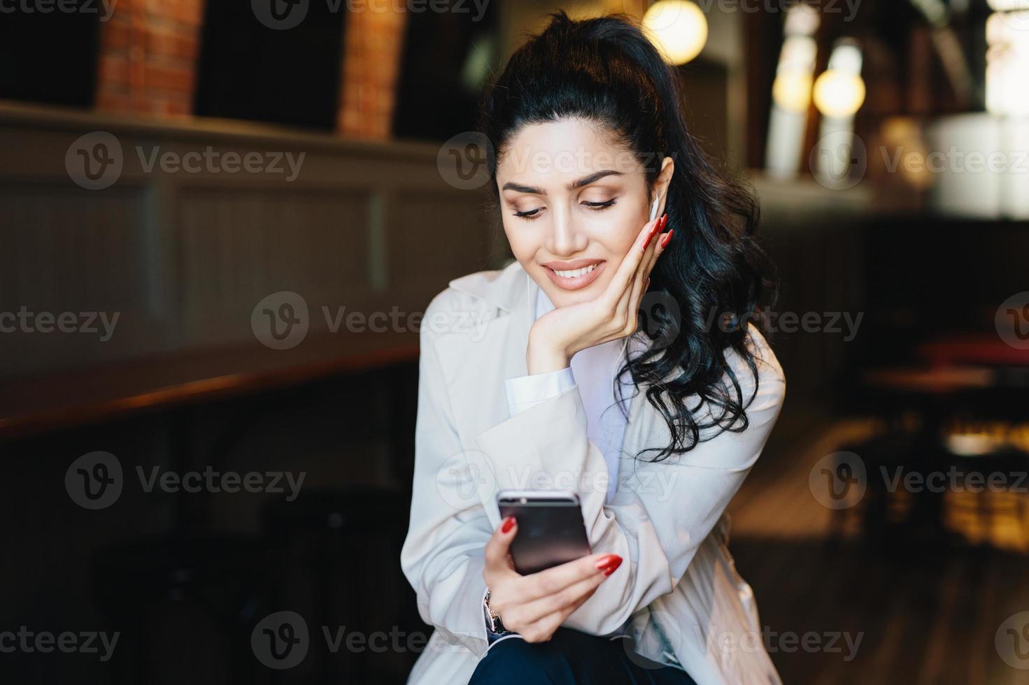 People, communication and lifestyle concept. Happy brunette woman with attractive face looking elegantly making video call, using online app on her mobile phone or listening to music relaxing indoors photo