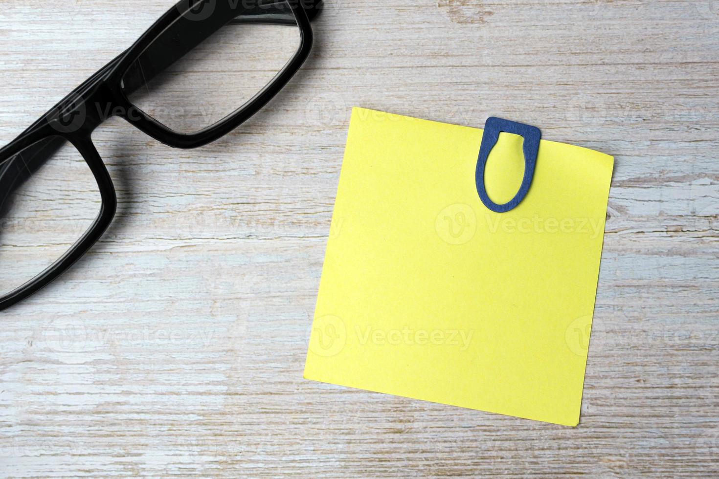 Yellow stick note with blue paperclip and reading glasses on wooden desk. photo