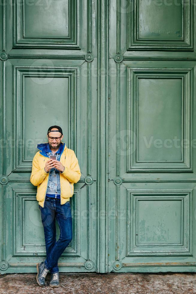 retrato completo de un hipster de moda con gorra de pie con las piernas cruzadas con un smartphone aislado sobre una antigua puerta verde. apuesto hombre barbudo leyendo sms en un teléfono celular genérico, enviando mensajes a amigos foto