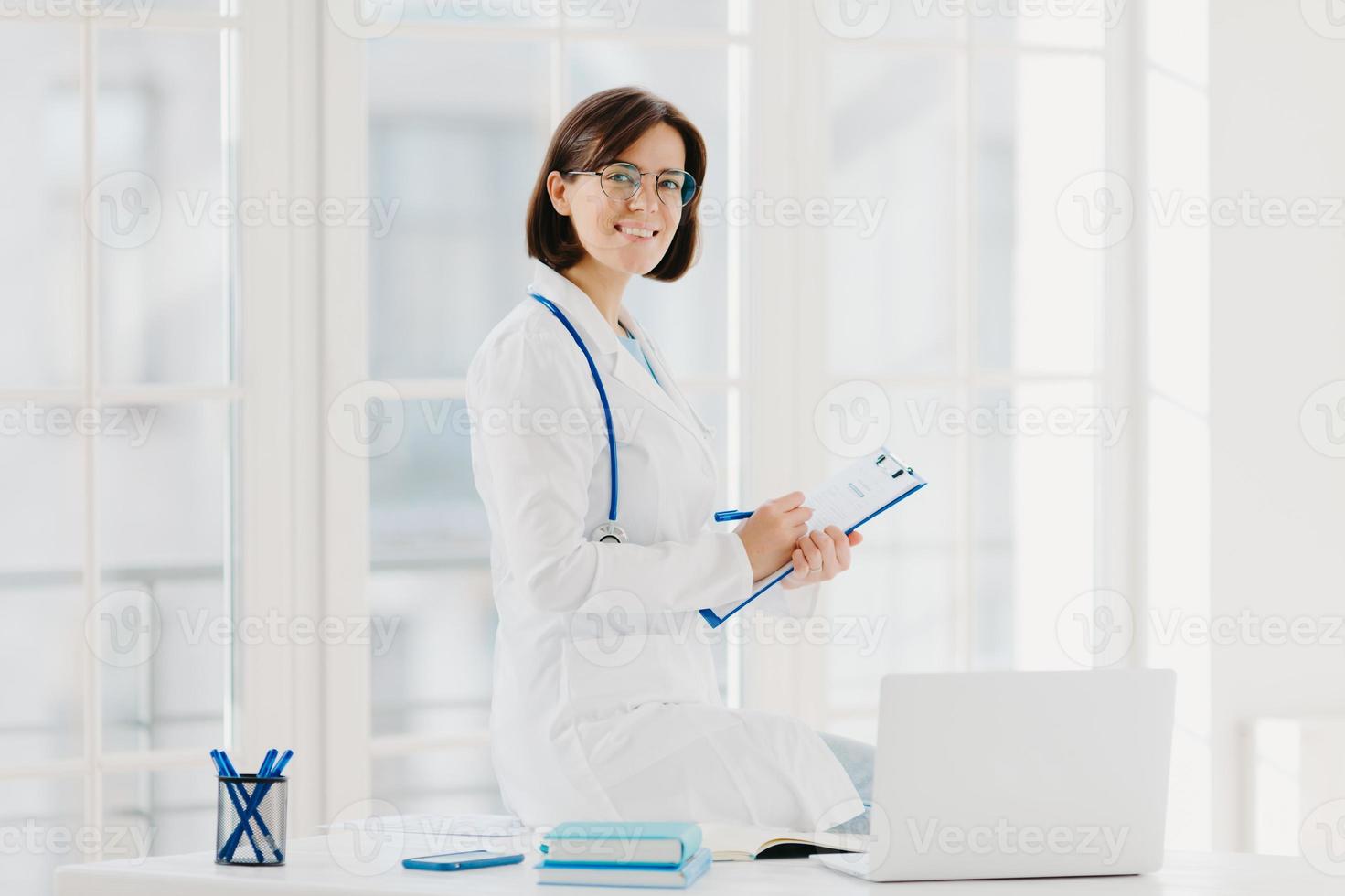 Woman doctor sits on table, writes down information in clipboard, poses at workplace in hospital office, works with laptop computer, smiles pleasantly. Health, technology, medicine, internet concept photo