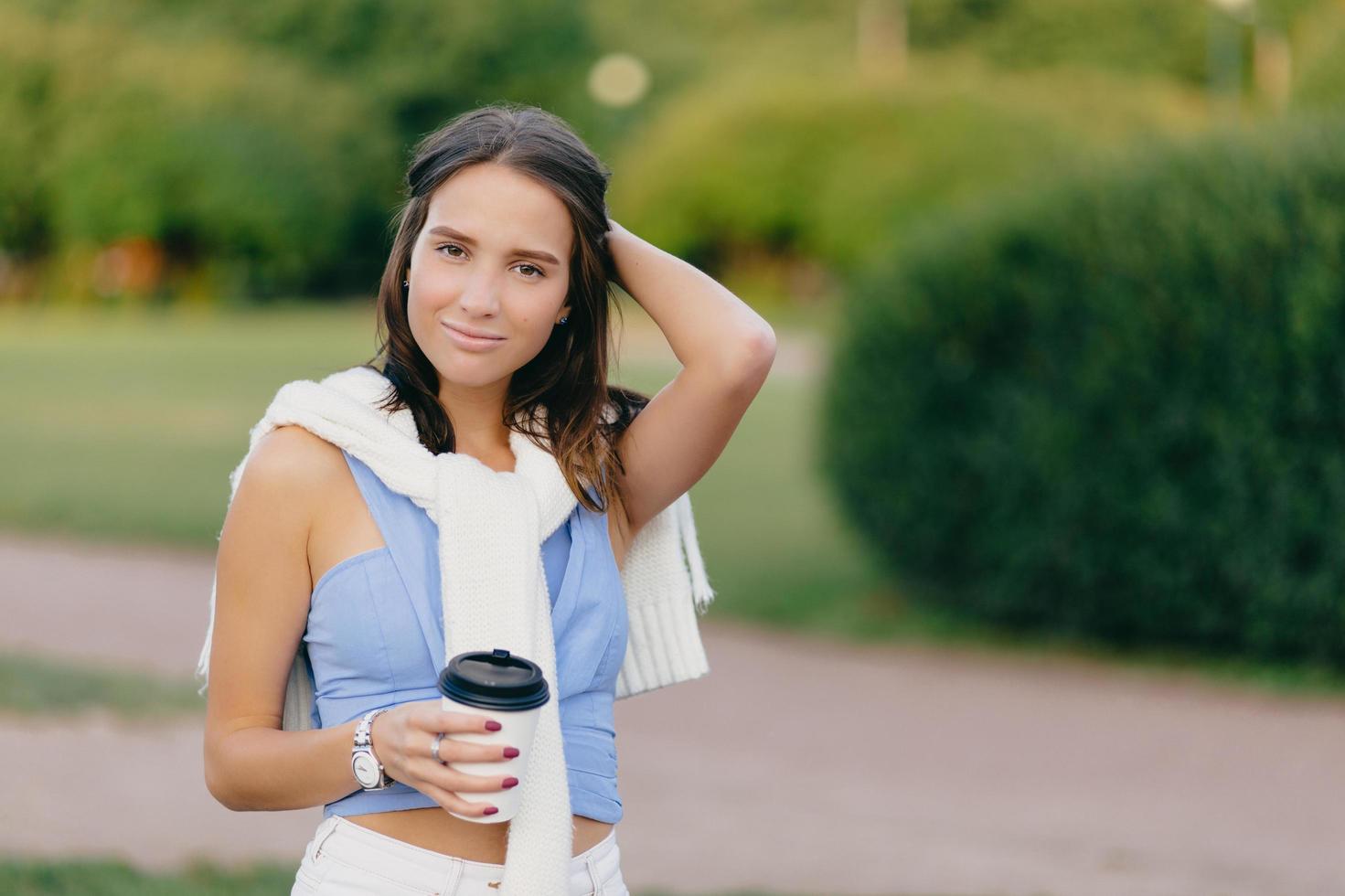 Pleased beautiful European woman with fit figure, keeps hand on head, drinks takeaway coffee, stands against green blurred background with copy space for your advertisement. Coffee time concept photo