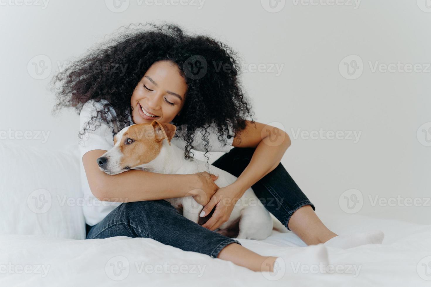 Pretty woman with curly hairstyle plays and enjoys with adorable small dog in bed, has good mood, pose together in cozy bedroom. Female wakes up with best friend. Lovely pet with owner indoor photo