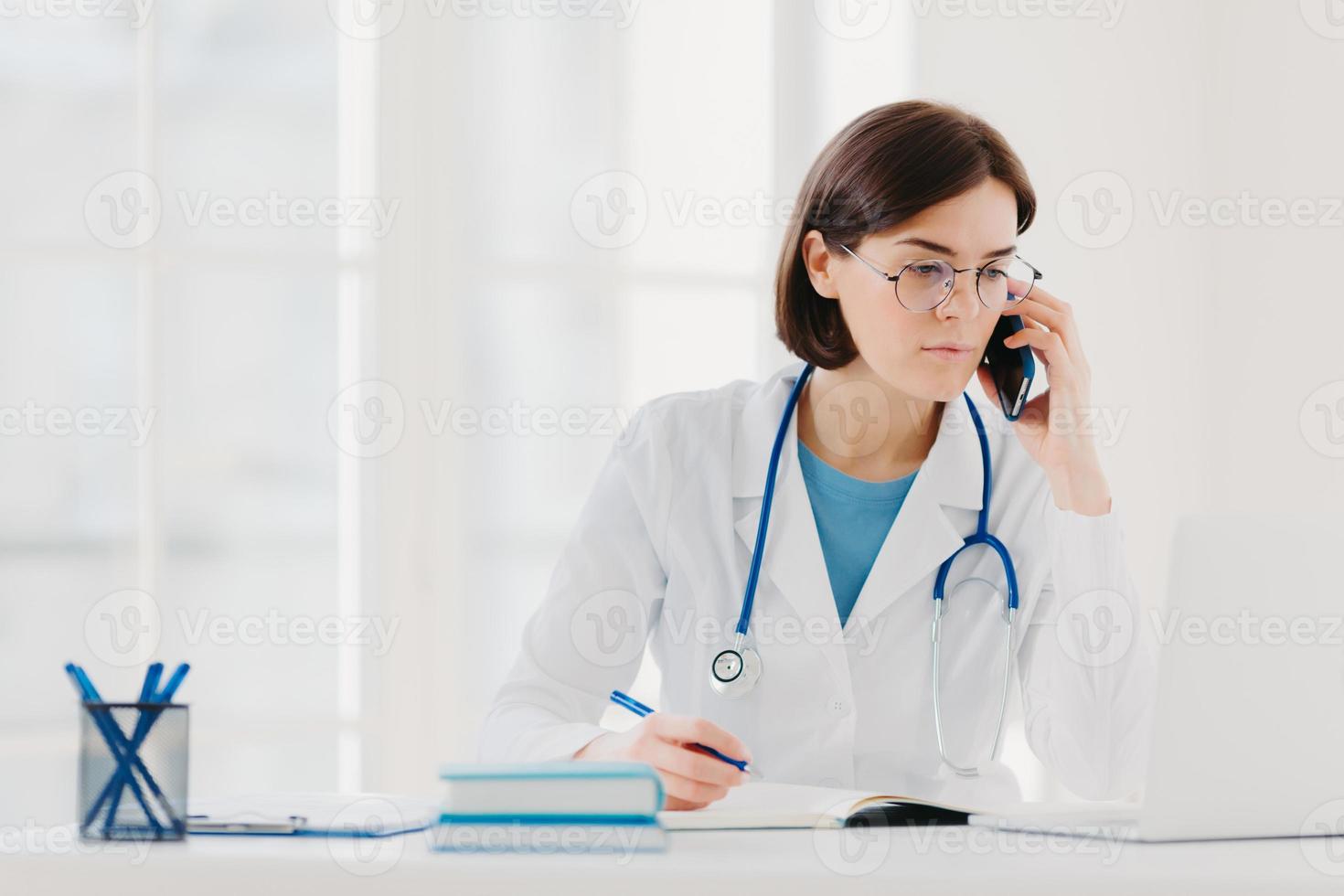 Heathcare personnel, medicine concept. Serious brunette female doctor focused at modern laptop computer, rewrites necessary information, talks on mobile phone, calls someone, has serious look photo