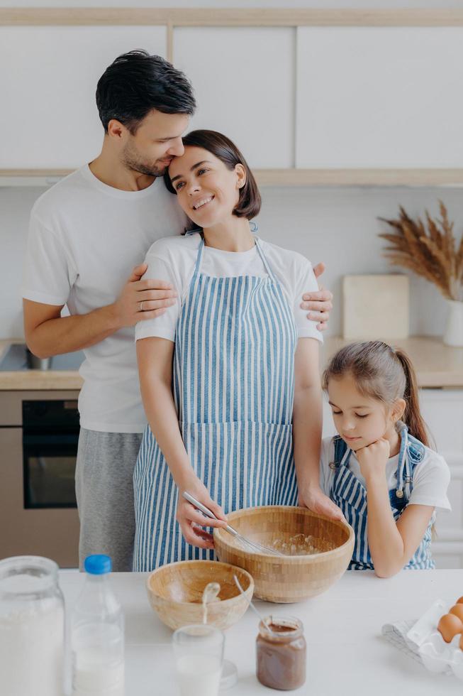 familia cocina comida en casa. el esposo y la esposa se abrazan con amor, la niña pequeña posa cerca, prepara una cena deliciosa, usa leche, chocolate, huevos, hace pasteles caseros, posa contra el interior de la cocina. foto