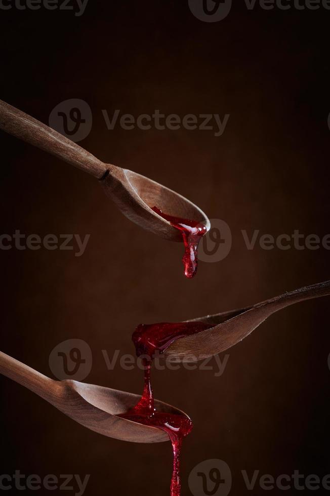 Berry red jam dripping from wooden spoon to spoon on brown background. photo