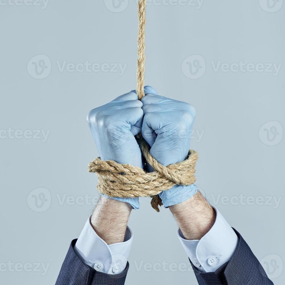 The businessman's hands in rubber gloves are tied with a rope and suspended on gray background. It is forbidden to work. photo
