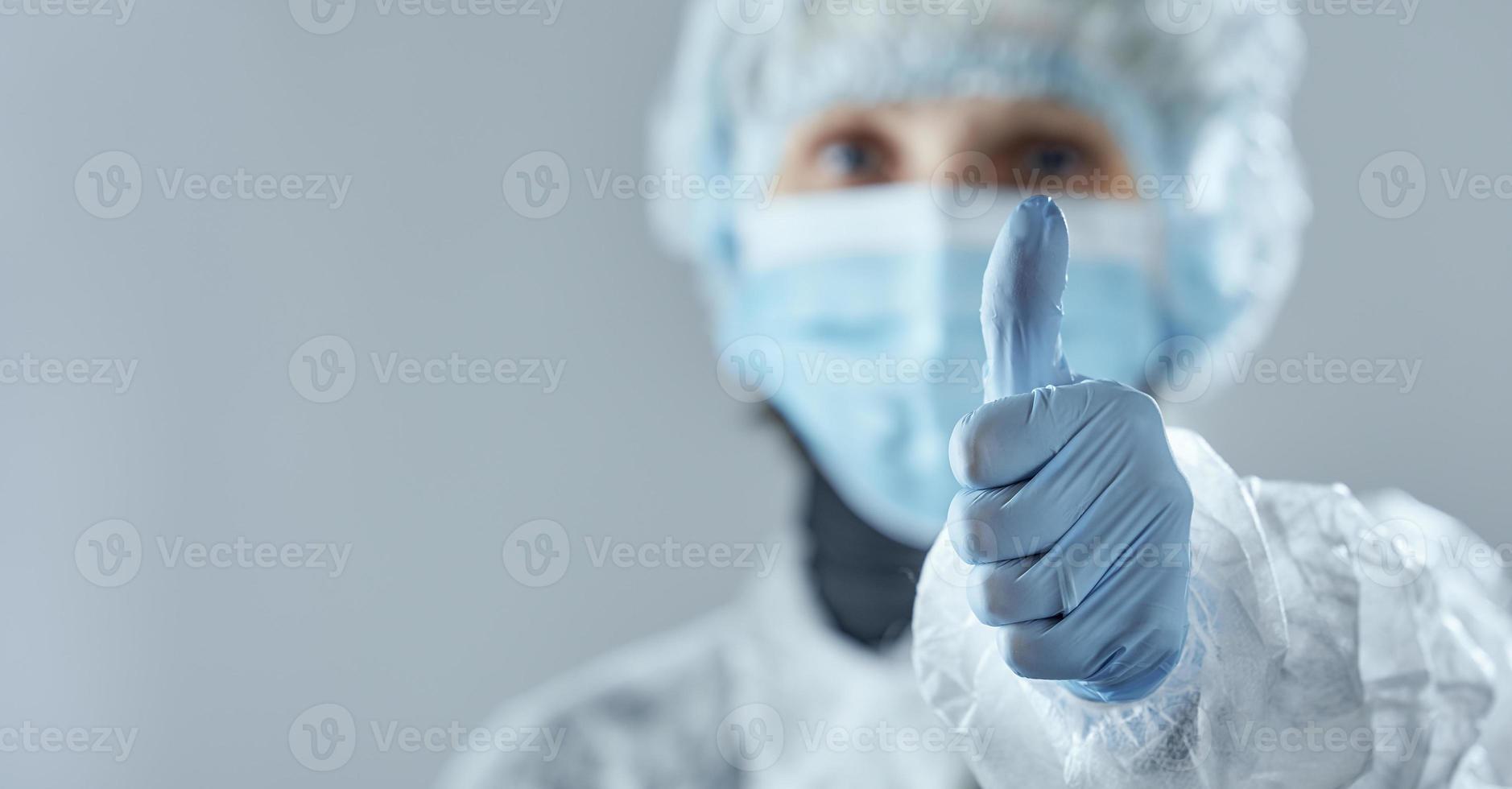 una mujer borrosa con una máscara médica, guantes de goma y ropa desechable muestra un signo de pulgar hacia arriba. foto con espacio de copia.