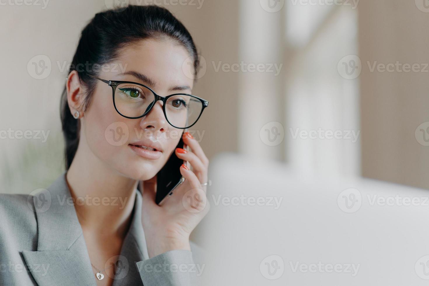 Close up shot of pleasant looking brunette European woman wears transparent glasses, focused in laptop screen, has telephone conversation during remote work, works on project. Technology concept photo