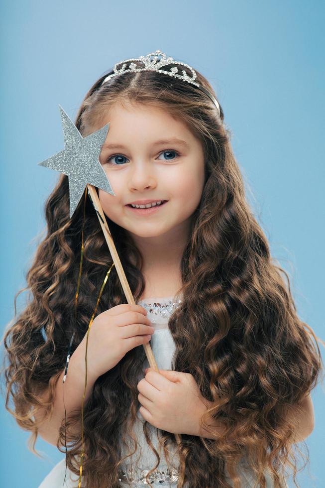 Image of attractive small child with curly dark hair, holds magic wand, looks happily into distance, beileves in miracle, isolated over blue background. Cute pretty little girl stands indoor photo