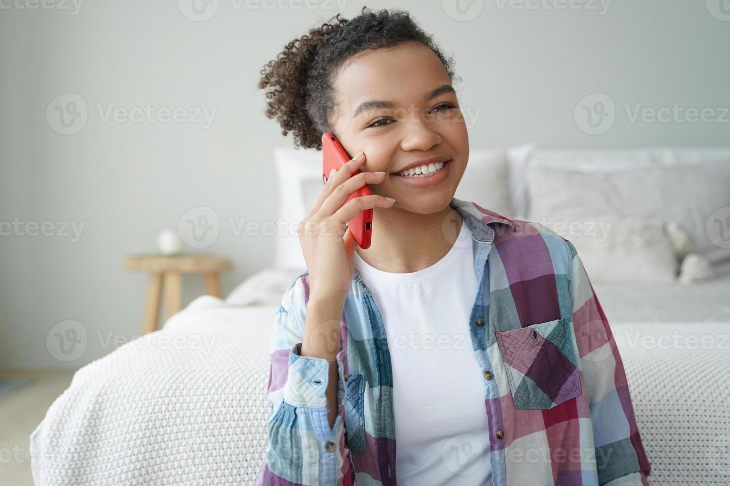 Smiling young african american teen girl making phone call, talking with friend in bedroom at