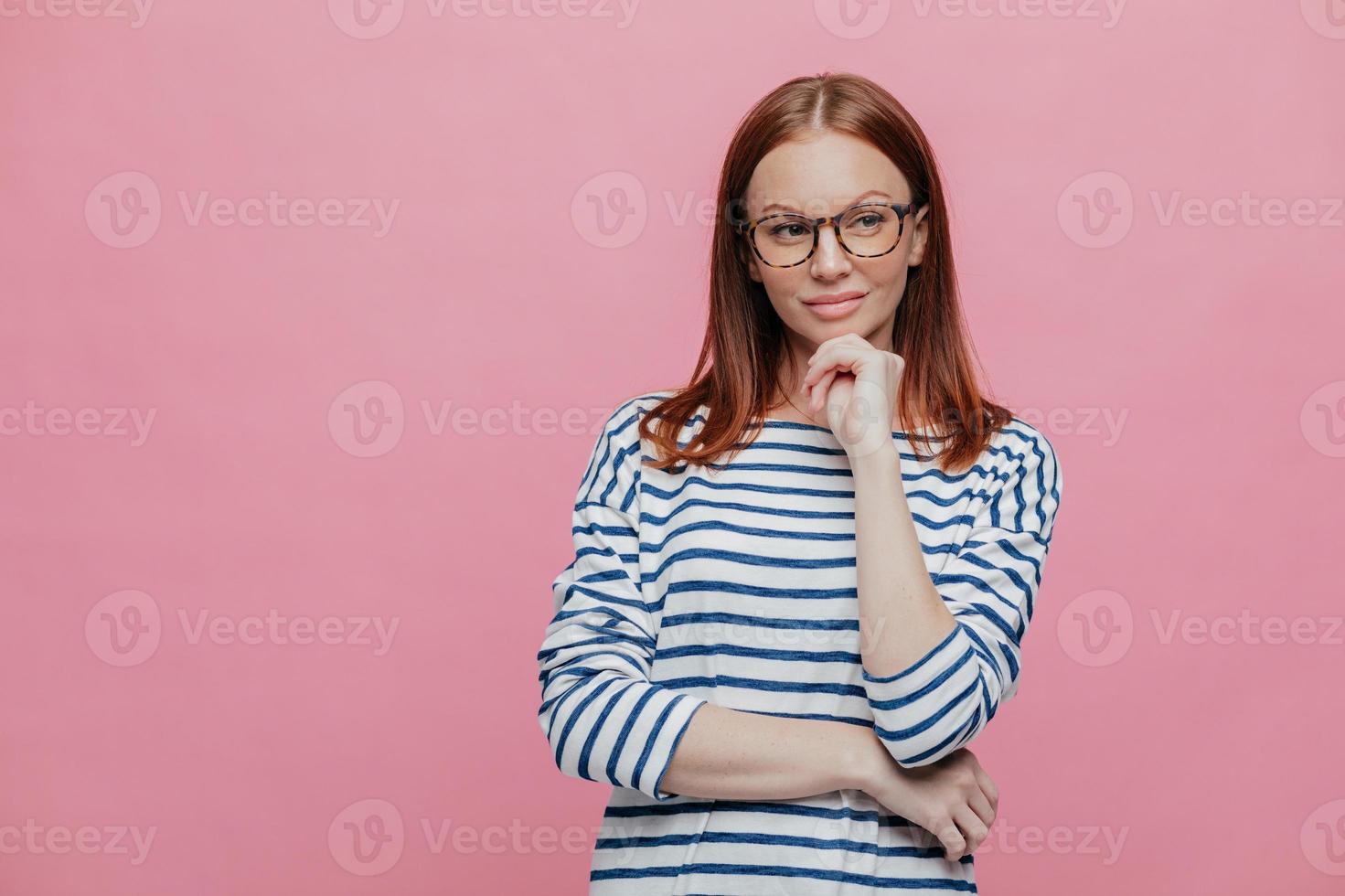 Waist up shot of dreamy thoughtful woman keeps hands partly crosed, holds chin, wears transparent glasses, looks pensively aside, models over pink background with copy space for your text or slogan photo
