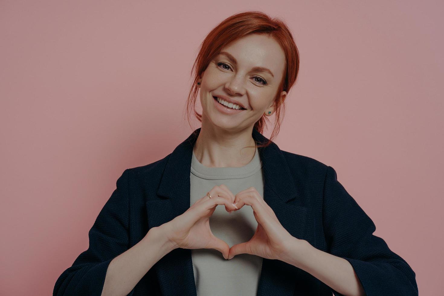 Isolated studio shot of young kind smiling redhead female making heart shape with both hands photo