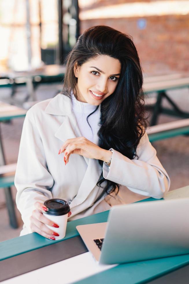 mujer joven con cabello oscuro con ojos brillantes, labios carnosos y piel sana con abrigo blanco descansando en el café y navegando por Internet usando una computadora portátil bebiendo café sabroso. concepto de belleza y juventud foto