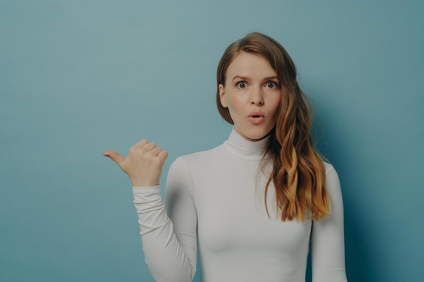 Excited millennial lady standing with thumb finger pointing aside on copy space for advertising product photo