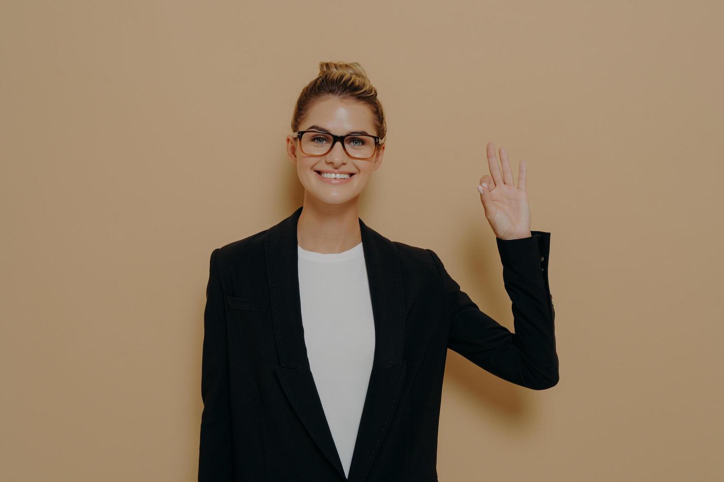 Positive young caucasian woman in classic wear isolated on beige background showing ok sign photo