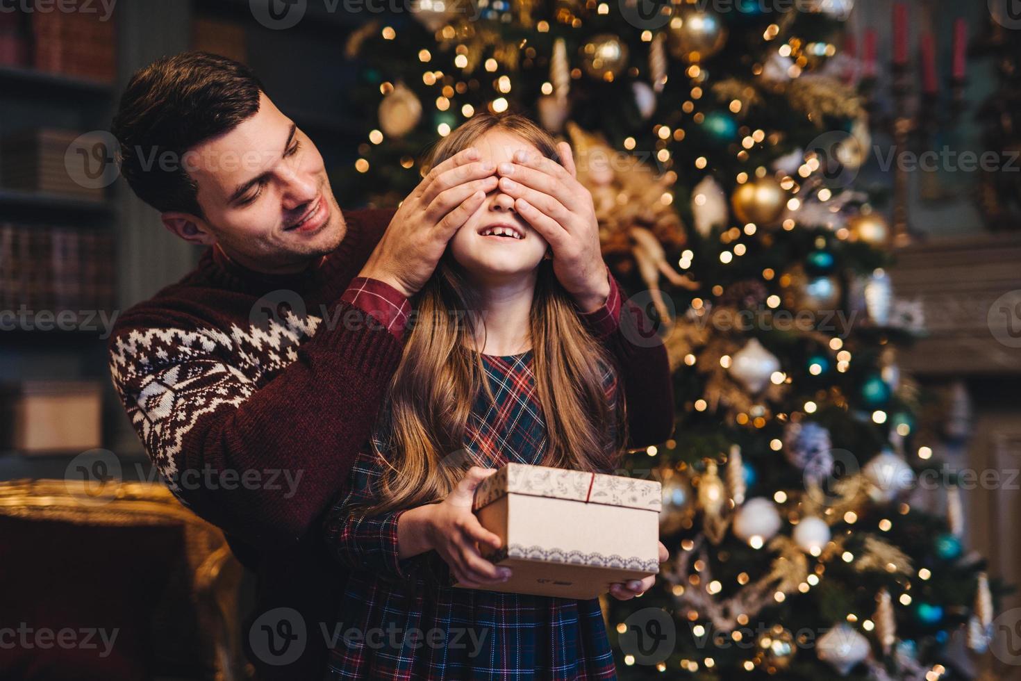 Portrait of young father covers his daughters eyes as going to make surprise for her, gives present, stand together near Christmas tree. Happy smiling girl recieves gift from dad. Surprise concept photo