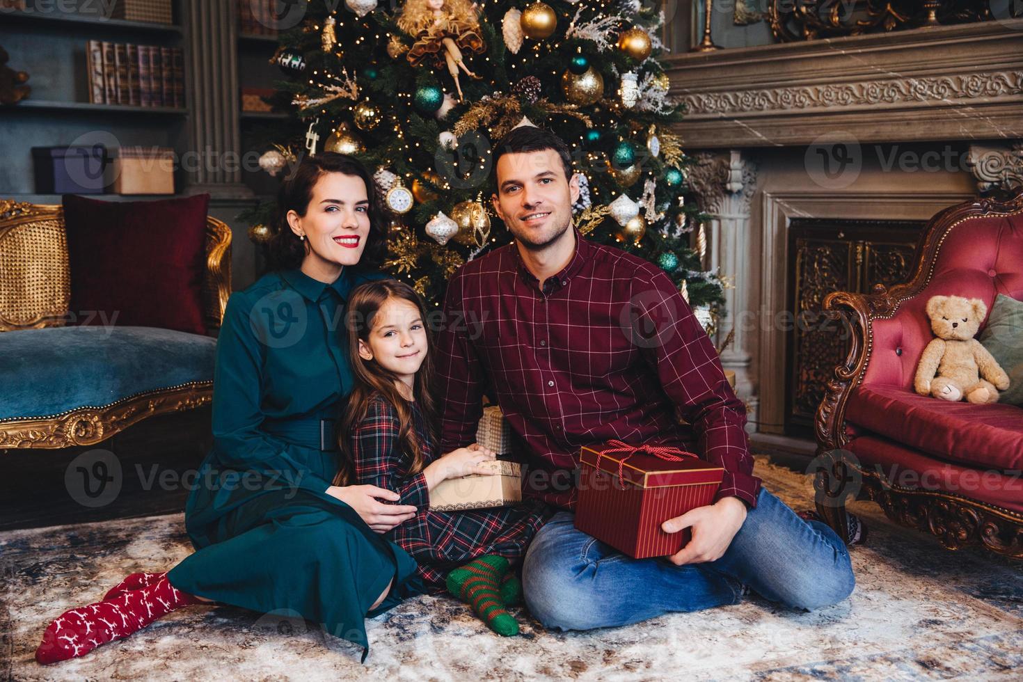 la foto interior de una familia joven tiene una buena relación, se sientan juntos en el piso cerca del árbol de año nuevo decorado, sostienen un regalo, se felicitan mutuamente por el próximo año nuevo y navidad. concepto de vacaciones