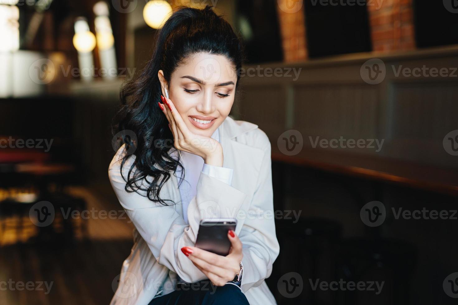 People, communication and lifestyle concept. Happy brunette woman with attractive face looking elegantly making video call, using online app on her mobile phone or listening to music relaxing indoors photo