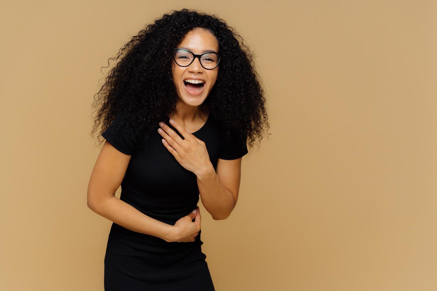 Beautiful dark skinned model cannot stop laughing, hears comic story, keeps hand on belly, laughs loudly, dressed in black clothes, stands against brown studio wall with empty space for information photo
