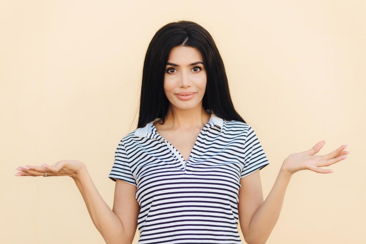 Adorable brunette young female with hesitant expression, shrugs shoulders in bewilderment, tries to find solution or makes decision, poses indoor alone. People and hesitation concept photo