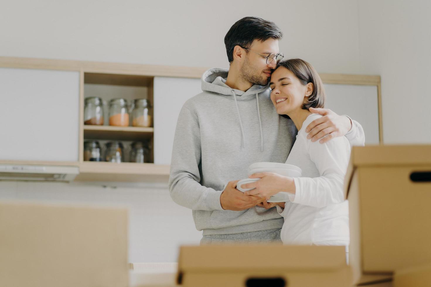 Young couple in love embrace and kiss with tender, hold pile of white plates, stand in kitchen during moving day, surrounded with many carton boxes filled with personal belongings, unpack stuff photo