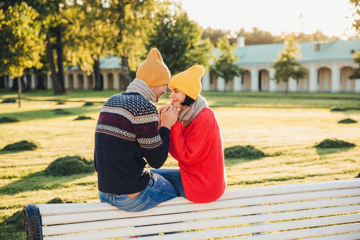 Romantic couple sit on bench, enjoy sunny day, keep hands together, look with great love at each other, have good relationships. Female has date with boyfriend in park, admire beautiful nature photo