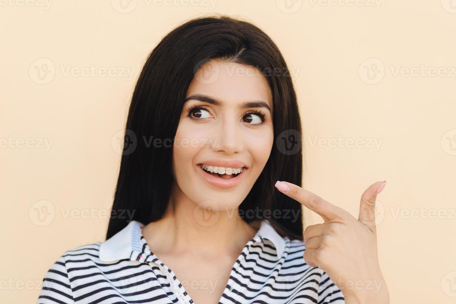 Pretty young brunette female with healthy skin, indicates at teeth with braces, wears striped t shirt, looks happily aside, shows her beauty, has make up, isolated over light studio background photo