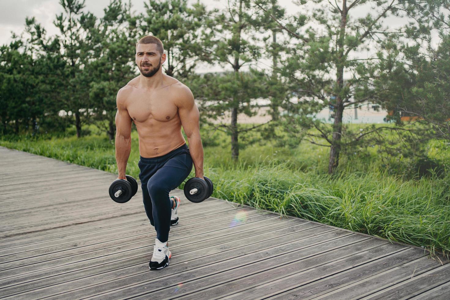 Motivated bearded man with naked torso makes squat with heavy barbells, does physical exercises outdoor, enjoys workout, poses near green trees, doing weightlifting. Healthy lifestyle concept photo