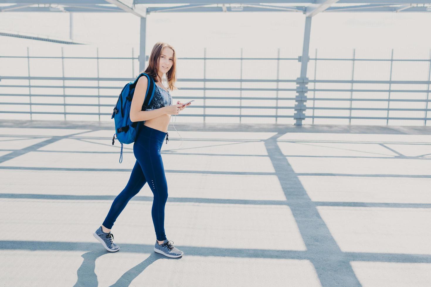 una linda joven camina afuera, lleva una mochila, sostiene un teléfono celular moderno, escucha música, revisa su canción favorita en la lista de reproducción, usa ropa deportiva informal y zapatillas de deporte, tiene un estilo de vida saludable foto