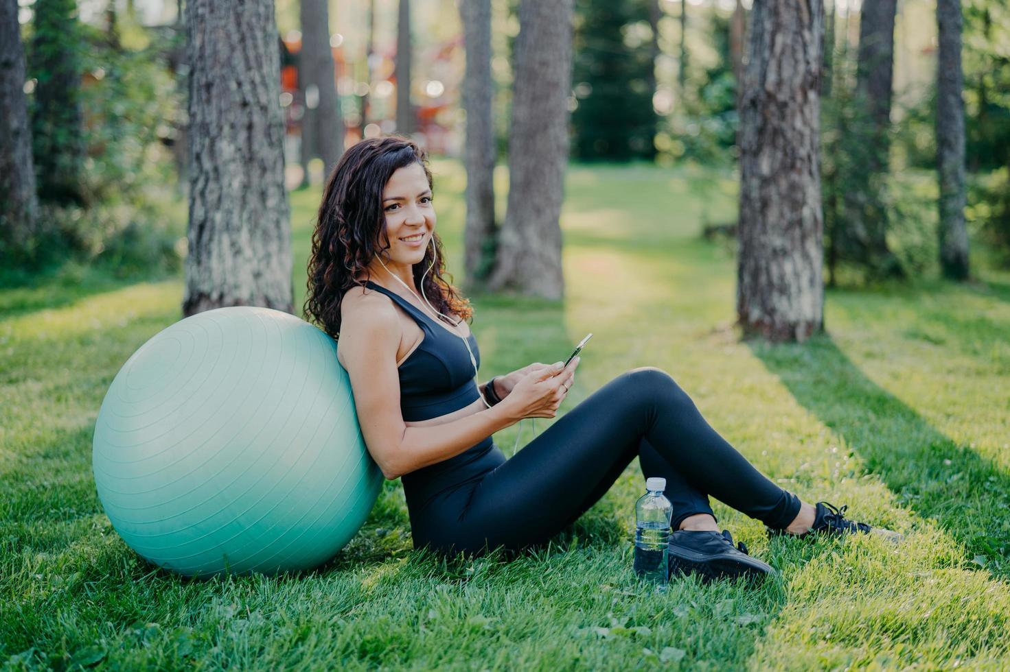 Photo of satisfied brunette young woman sits back to big fitness ball, uses mobile phone for listening music, poses on green grass in forest during sunny day, wears sports clothes. Sport concept