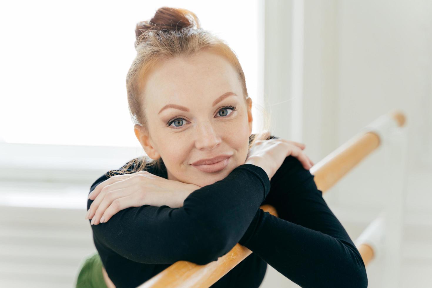 Beautiful young red haired female dancer crosses hands and leans on ballet barre which supports her during exercises practices dance in studio does stretching, being flexible looks directly at camera. photo