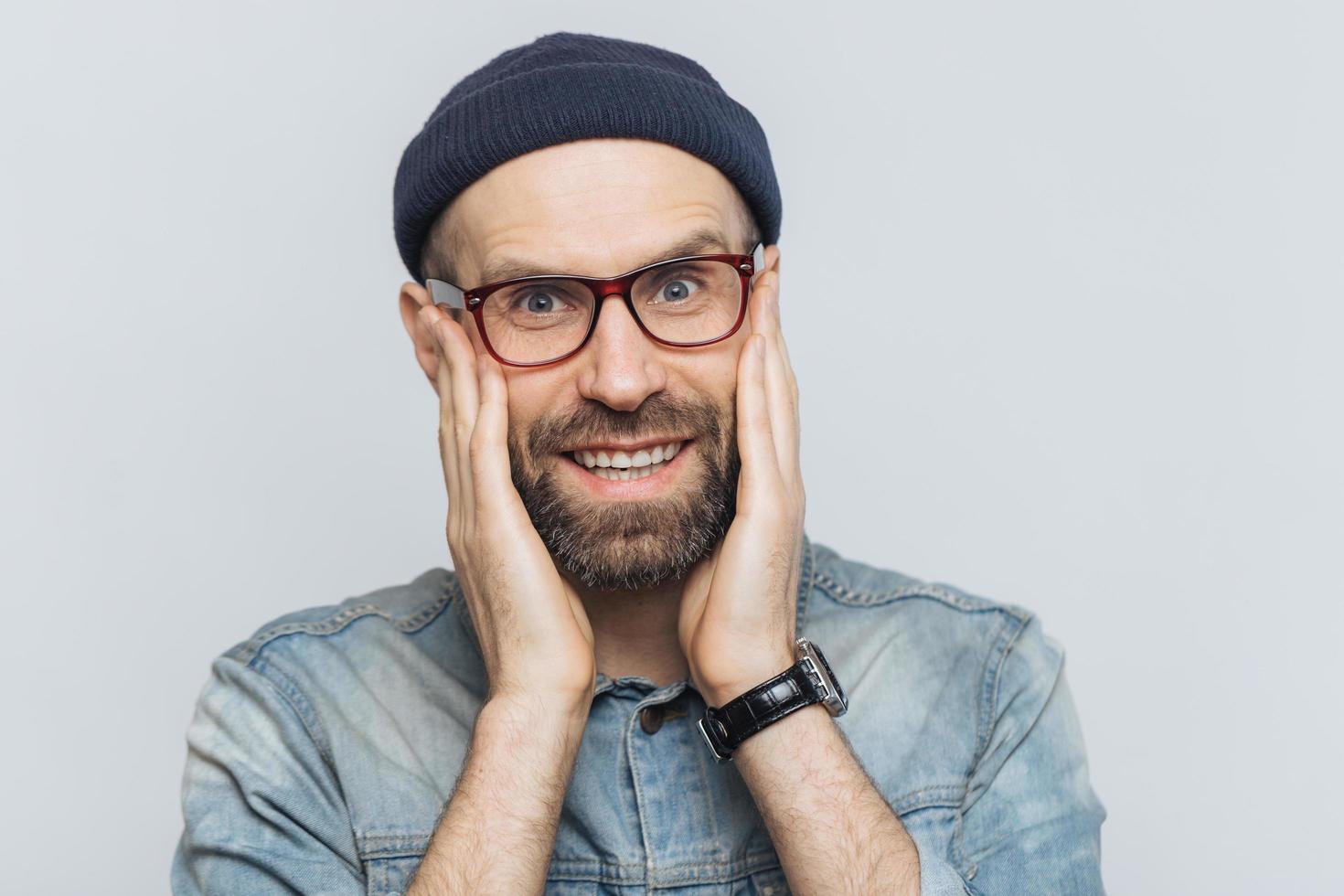 Portrait of attractive young man with thick beard and mustache looks with delighted expression, has white perfect teeth, poses in studio against blank copy space. People and positive emotions photo