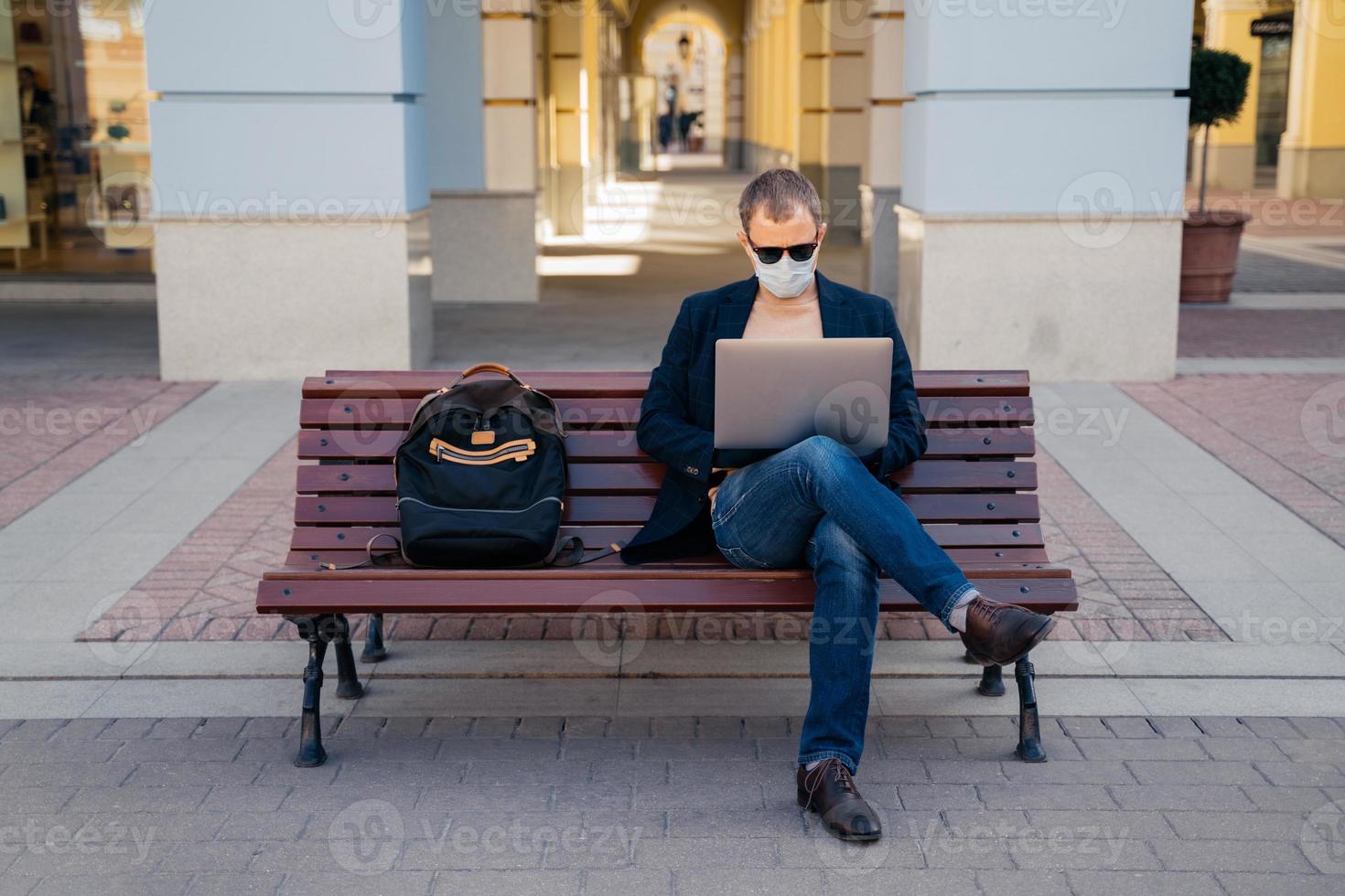 Photo of busy man freelancer in mask does distance work, keyborads on laptop computer, sits at wooden bench outside, avoids crowded places during quarantine time. Traveling and coronavirus concept