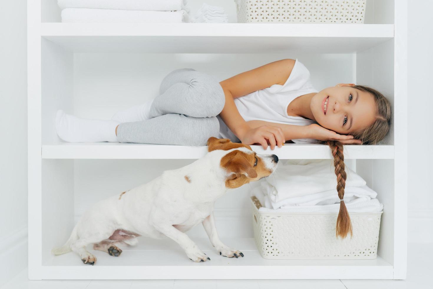 Photo of lovely preschooler lies on white shelf in washing room, has long pigtail, plays with dog, wears casual clothing, smiles gently, spends weekend at home. Children, animals and household concept