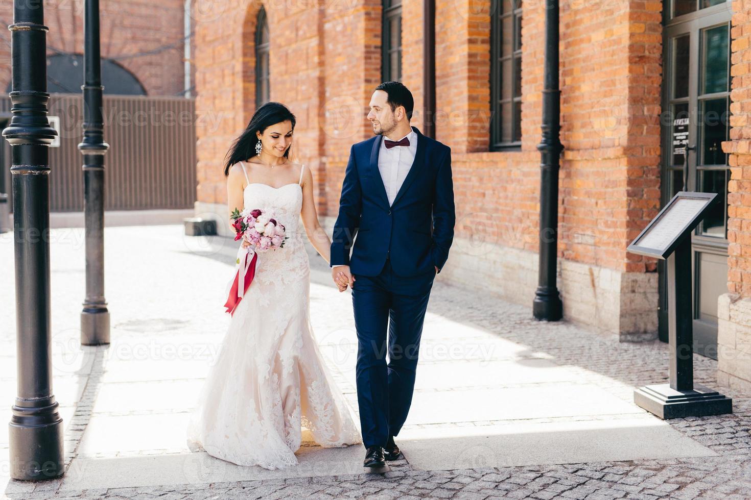 Horizontal portrait of affectionate lovely married couple, keeps hands together, walk outdoor, look at each other with love and passion. Bride in wedding dress and bridegroom in black formal suit photo