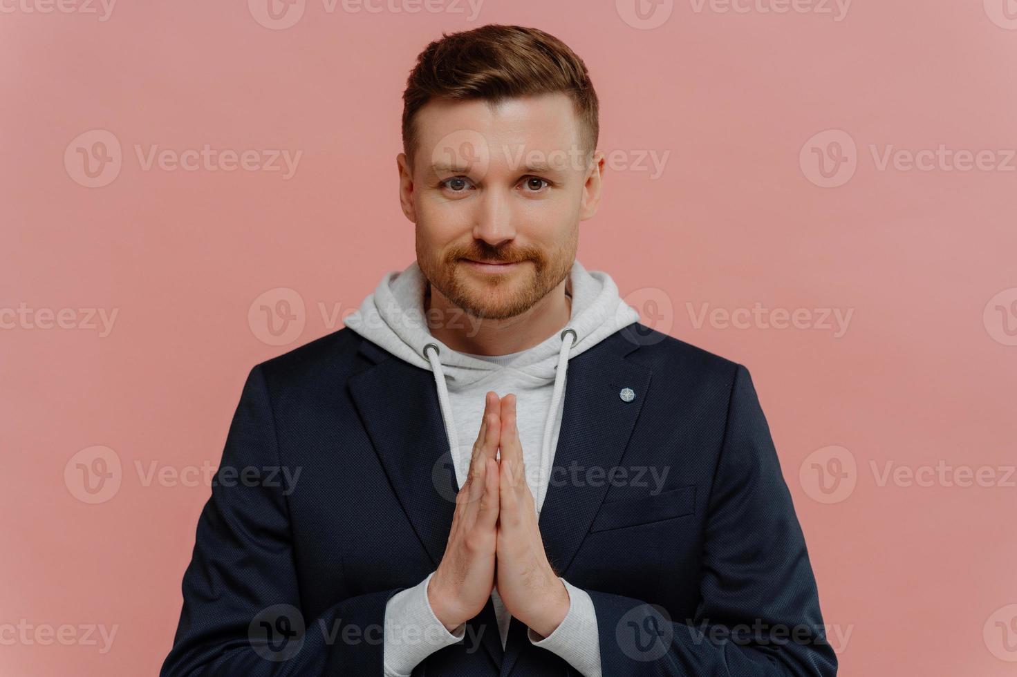 Serious bearded adult man keeps palms pressed together prays for something looks directly at camera believes in something good happen poses against pink background. Handsome guy makes request photo