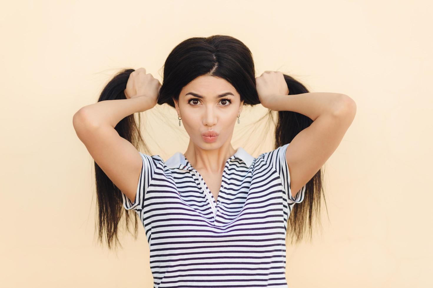 Portrait of pretty brunette female model makes two pony tails from her luxurious hair, has dark eyes, keeps lips round, wears casual t shirt, isolated over light studio background. People and fashion photo
