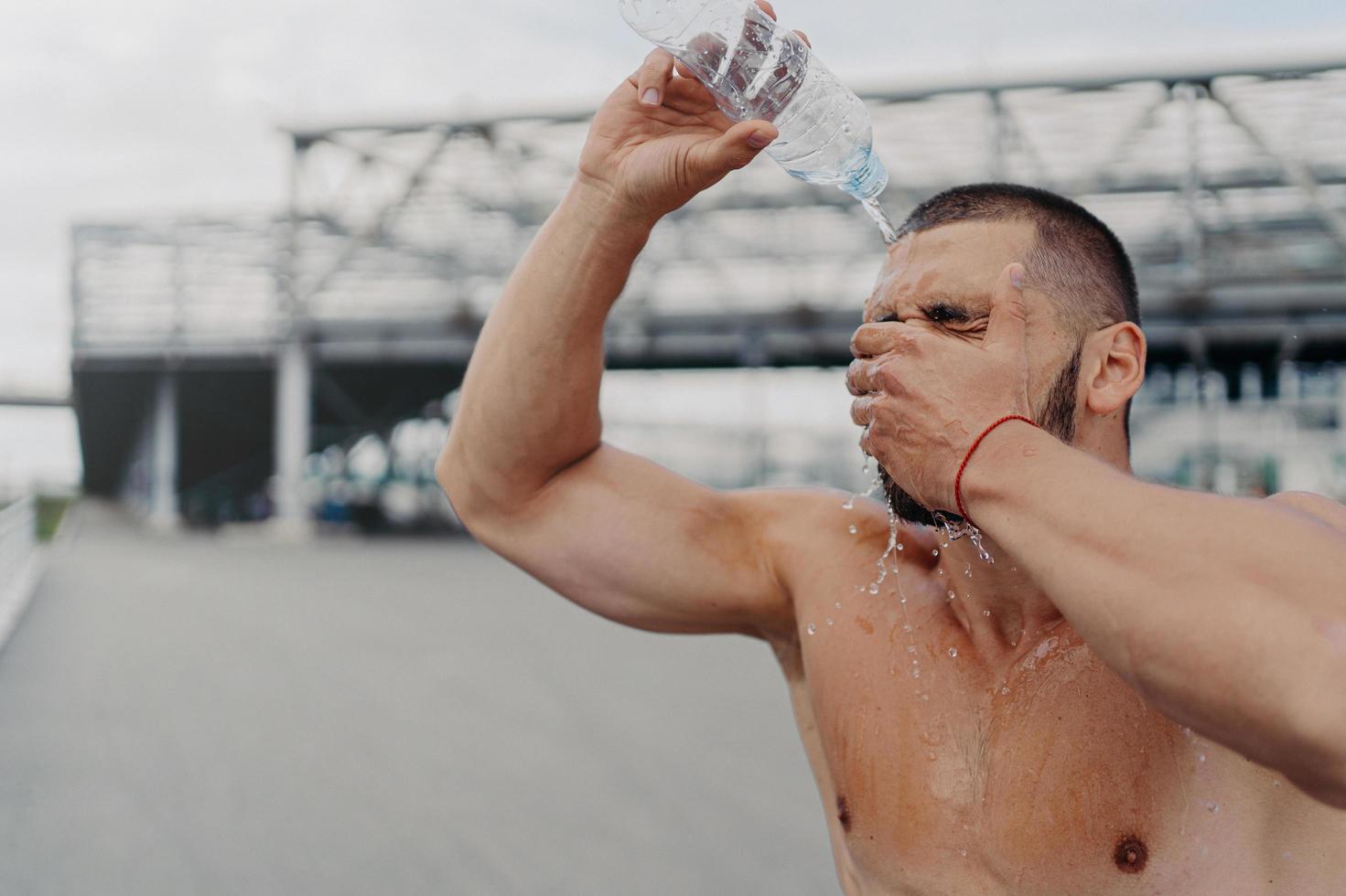 Image of athletic guy with muscular body pours water on himself from bottle, recovers after hard workout, feels tired and thirsty, cools body with cold liquid, loves sport. Healthy lifestyle photo