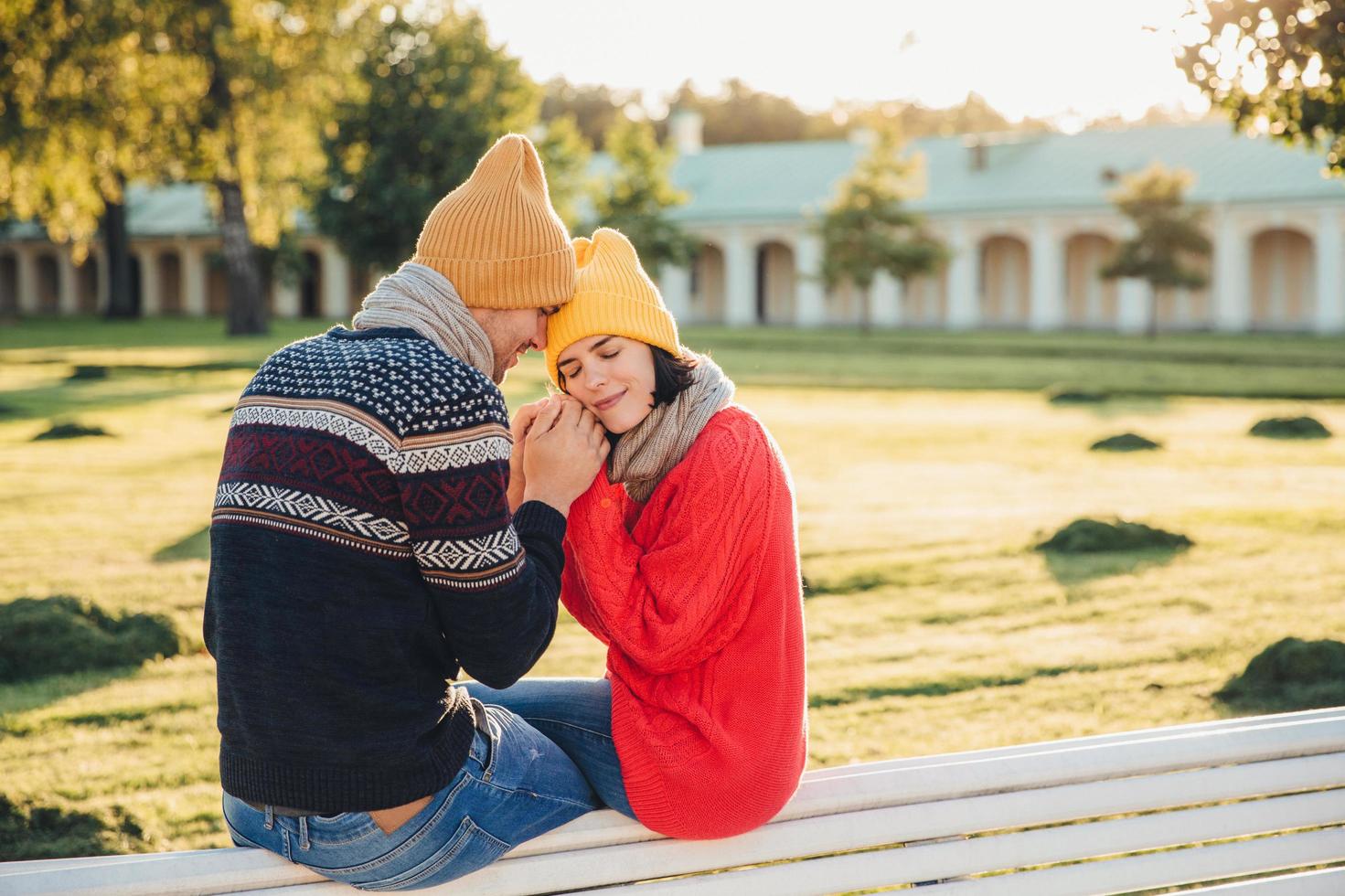 True feelings and romantisism concept. Adorable young woman in knitted yellow hat and red warm sweater warms her hands in boyfriends hands, sit together on bench, closes eyes with great enjoyment photo