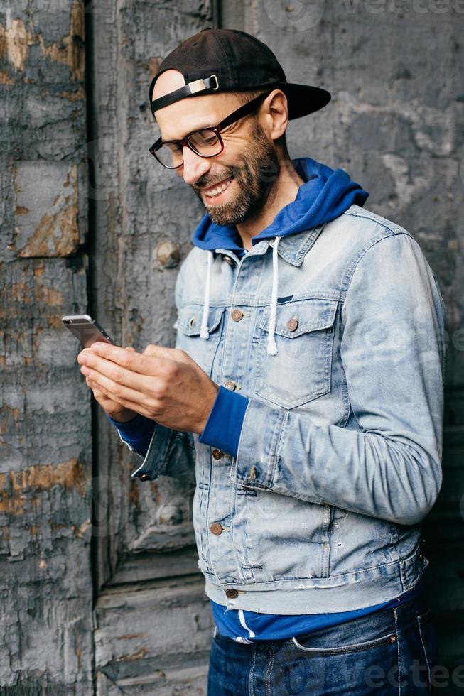 Vertical portrait of hipster guy in denim shirt, cap and eyewear holding modern telephone in his hands having glad expression while communicating with his girlfriend. Lifestyle and technolgy concept photo