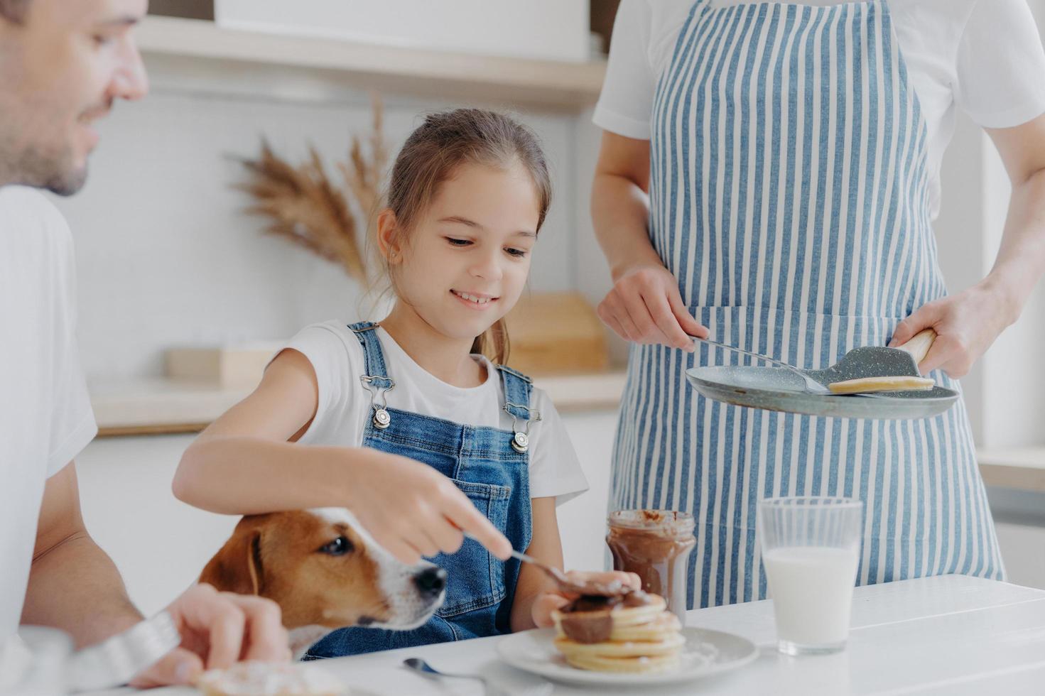 la pequeña niña feliz disfruta comiendo un delicioso postre preparado por mamá, agrega chocolate derretido a los panqueques, disfruta estar juntos y madre, padre y perro, toman un delicioso desayuno nutritivo en la cocina foto