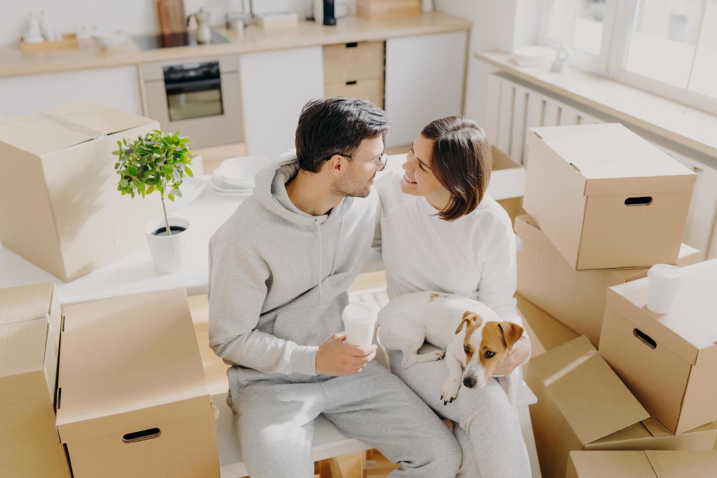 una pareja sonriente se expresa buenos sentimientos románticos, acaba de llegar a una casa nueva, posa alrededor de contenedores de cartón, bebe café para llevar, muebles de cocina en el fondo, perro en manos de mujeres foto