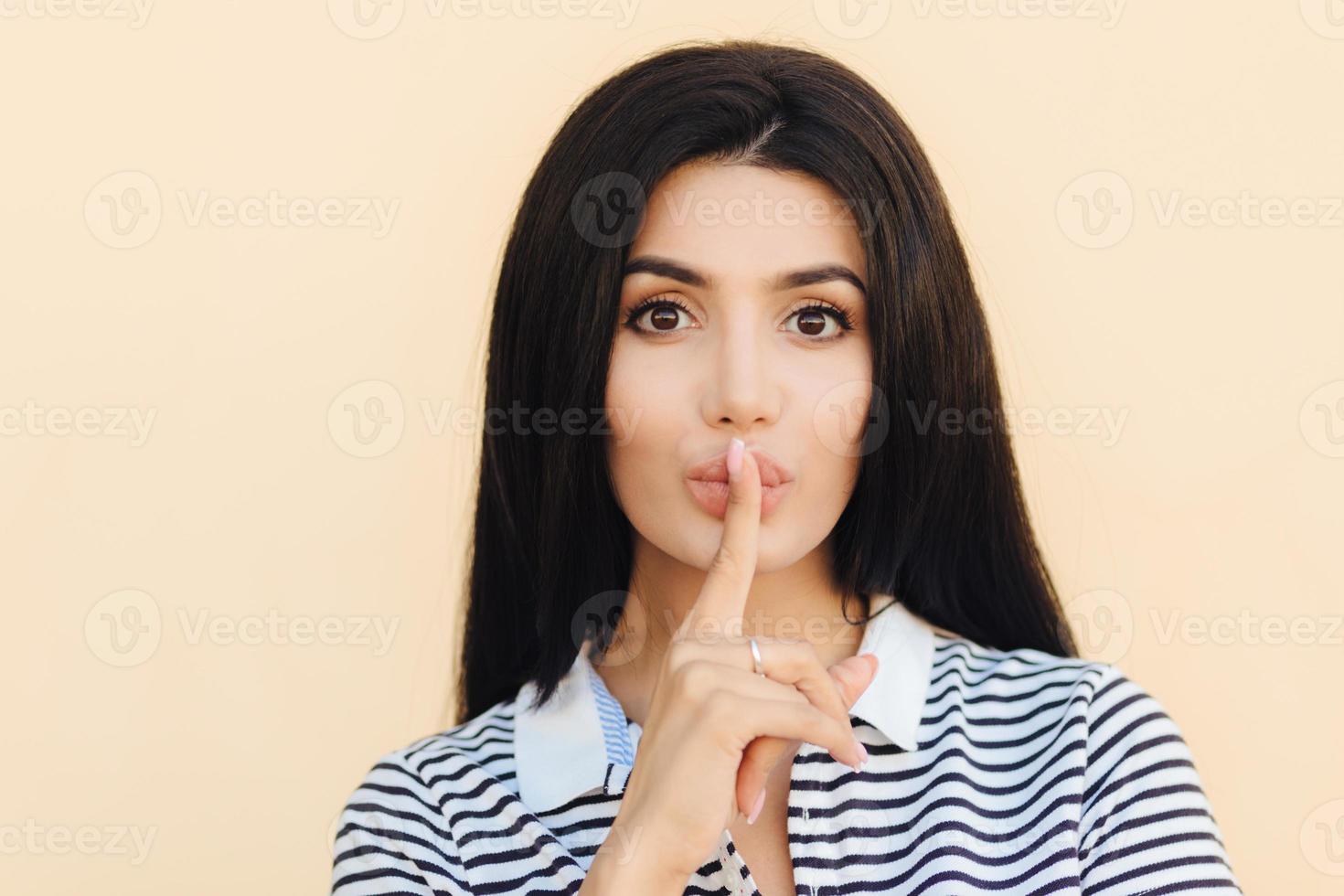 People and hush concept. Attractive female with silence sign keeps fore finger on lips, asks to be quiet as someone is sleeping, dressed in casual clothes, gestures indoor. People and conspiracy photo