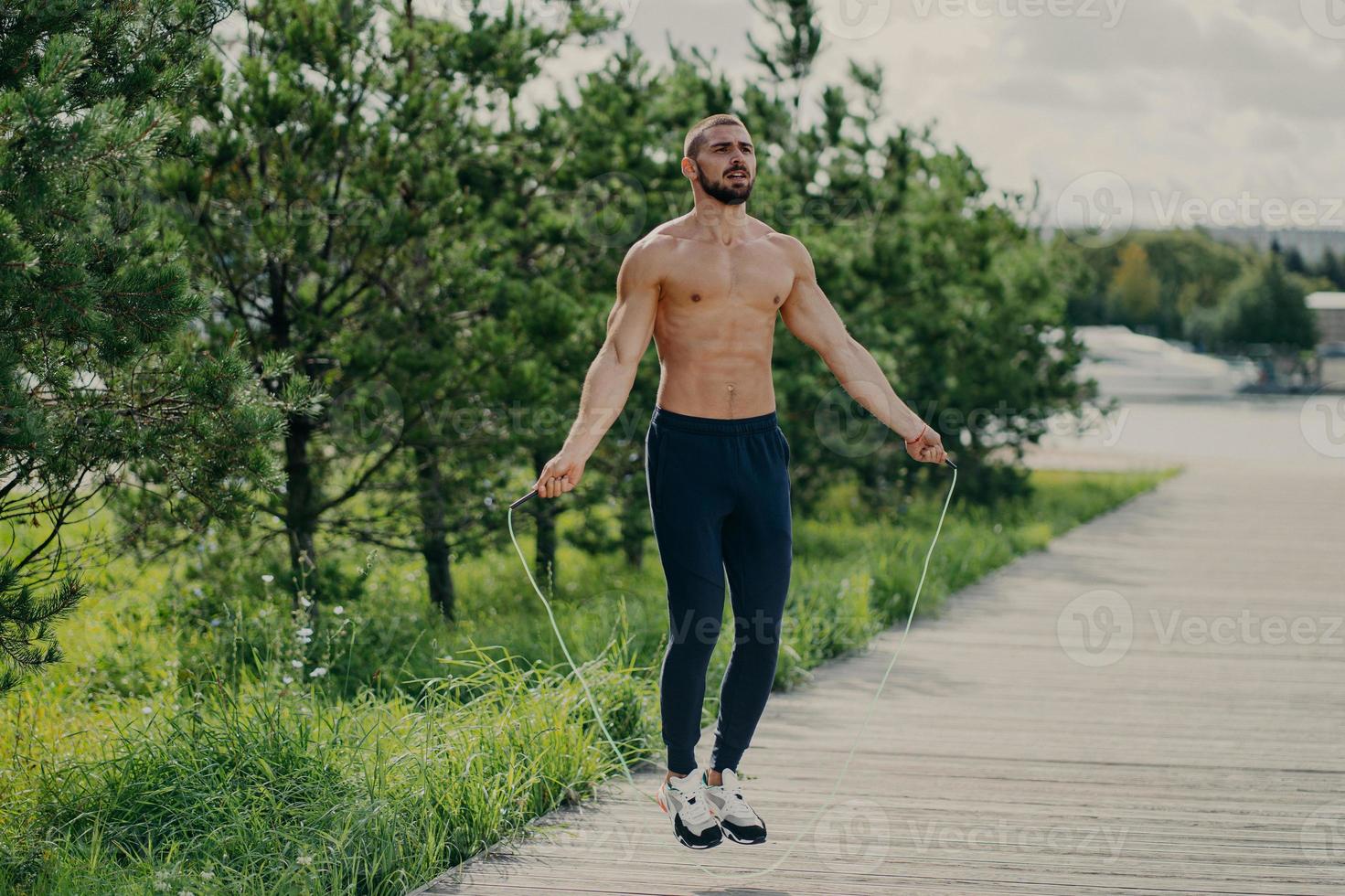 Sporty man with thick beard does exercises with jumping rope, has muscular torso, stays in good physical shape, poses outdoor. Fitness and healthy concept. Male runner warms up with sport equipment photo